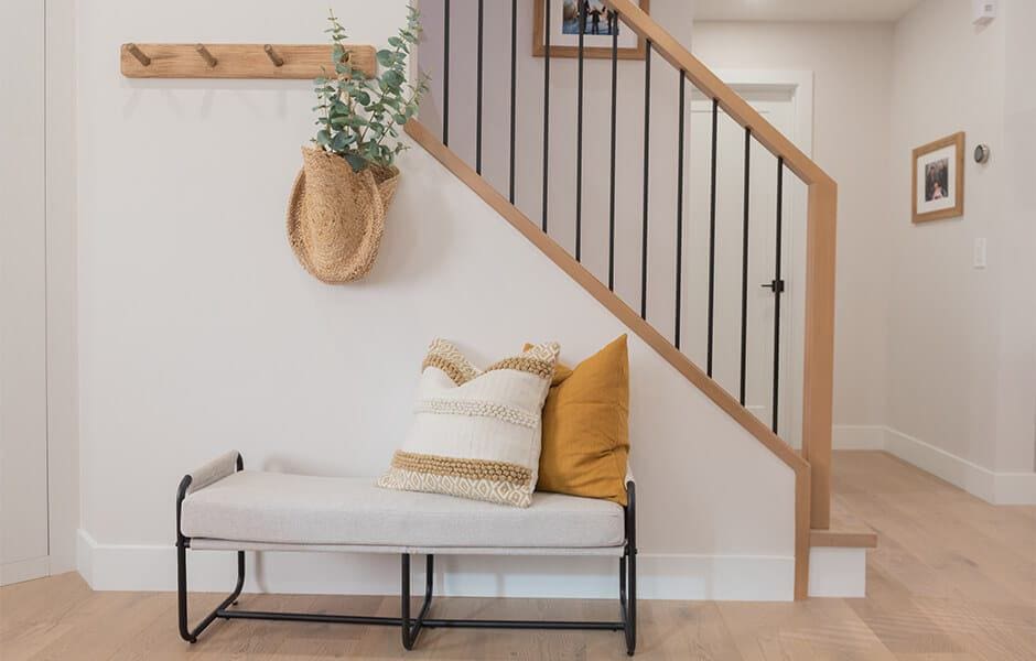 An entryway with a white wall, a wooden staircase with black railings, and a white bench with yellow and white pillows