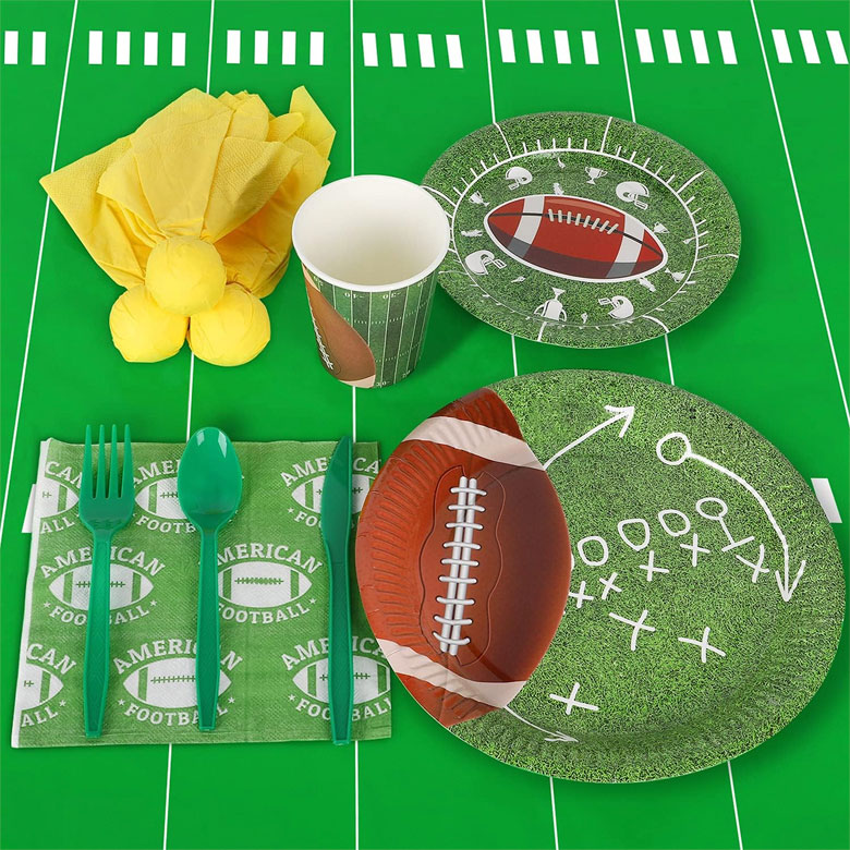 Football-themed party tableware with plates, cup, napkins, and utensils on a green field tablecloth