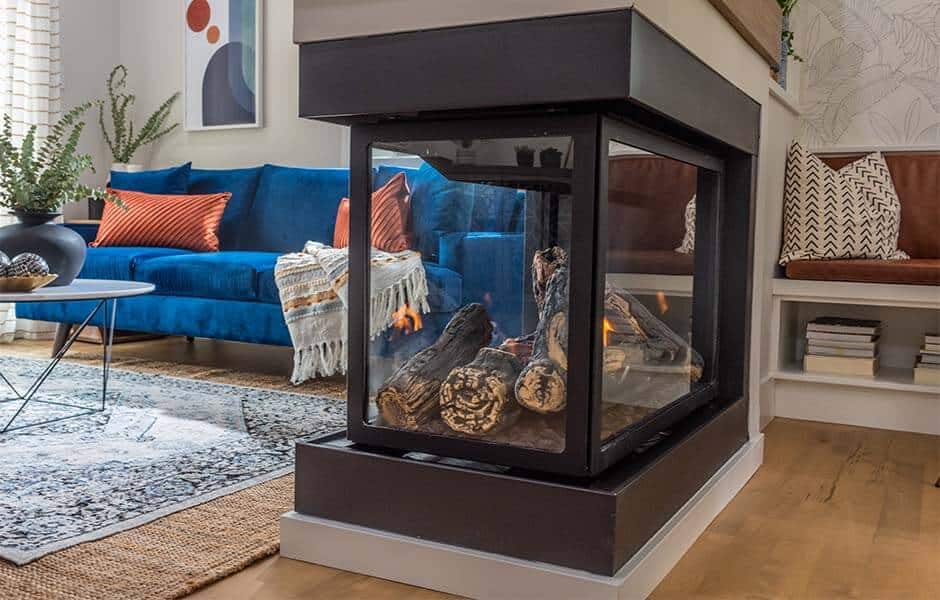 A three-sided fireplace, blue sofa with orange pillows, coffee table, patterned rug, and wood logs