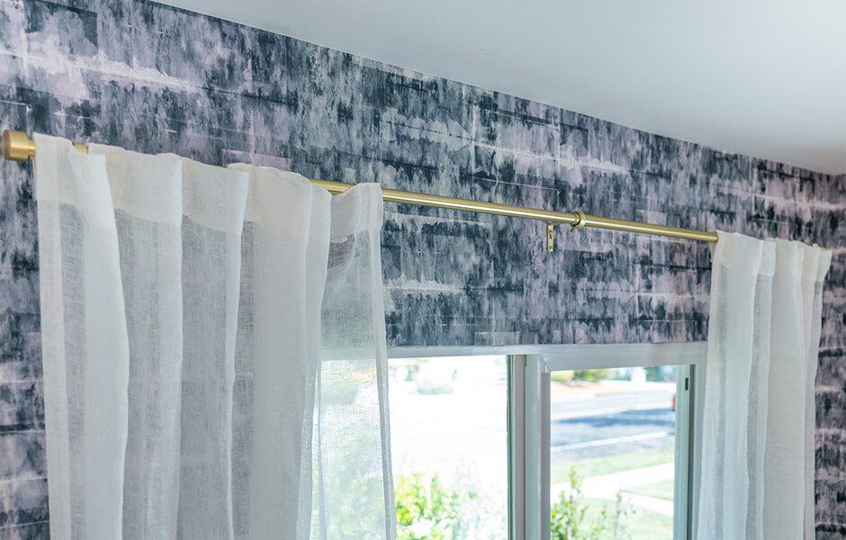 Gold curtain rod with sheer white curtains against a dark textured wall above a window