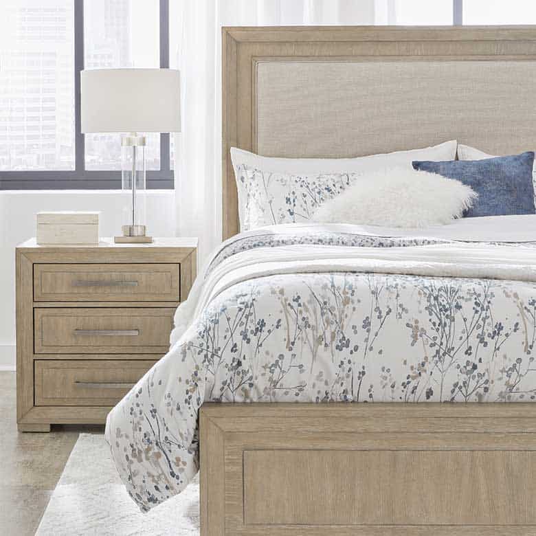 Light wood bedroom set with floral bedding, white and blue accent pillows, and a clear glass table lamp