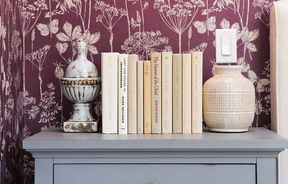 Gray dresser with neutral books, ceramic urn, and vase against purple floral wallpaper