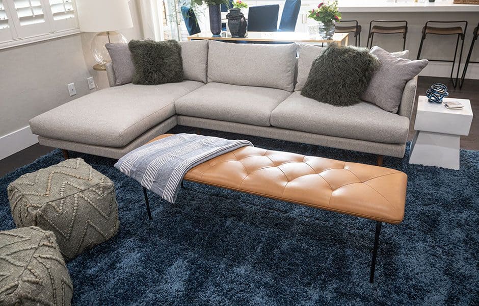 Living room with a gray sectional sofa, leather bench, blue rug, and textured poufs