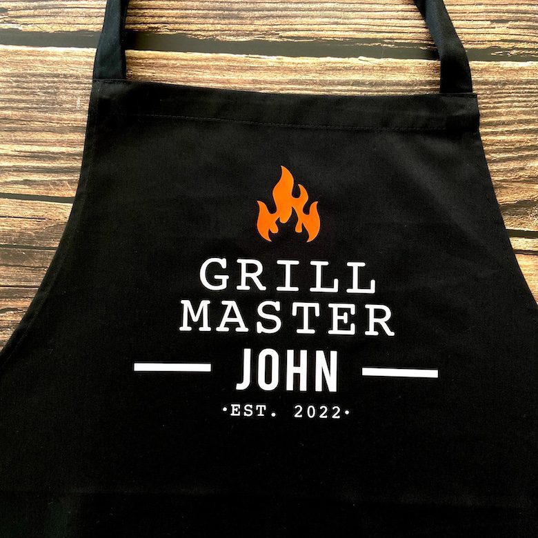 Black apron with "Grill Master John" and "Est. 2022" printed beneath an orange flame graphic