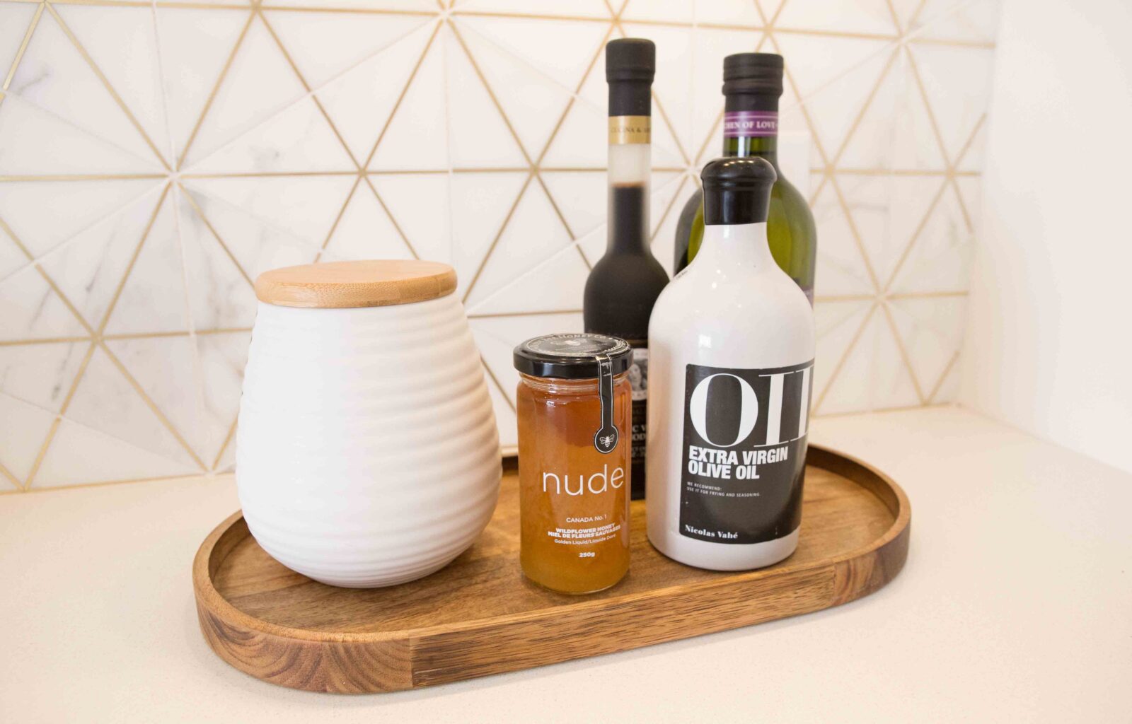 Olive oil, honey jar, and white canister on a wooden tray with geometric tile background