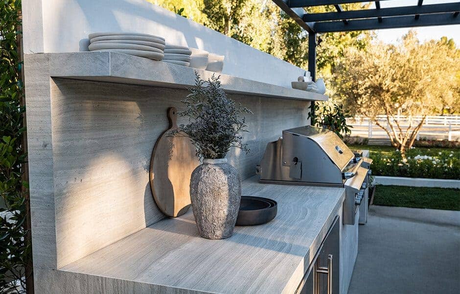 Outdoor kitchen with built-in grill, shelves with dishes, and a decorative vase on the counter