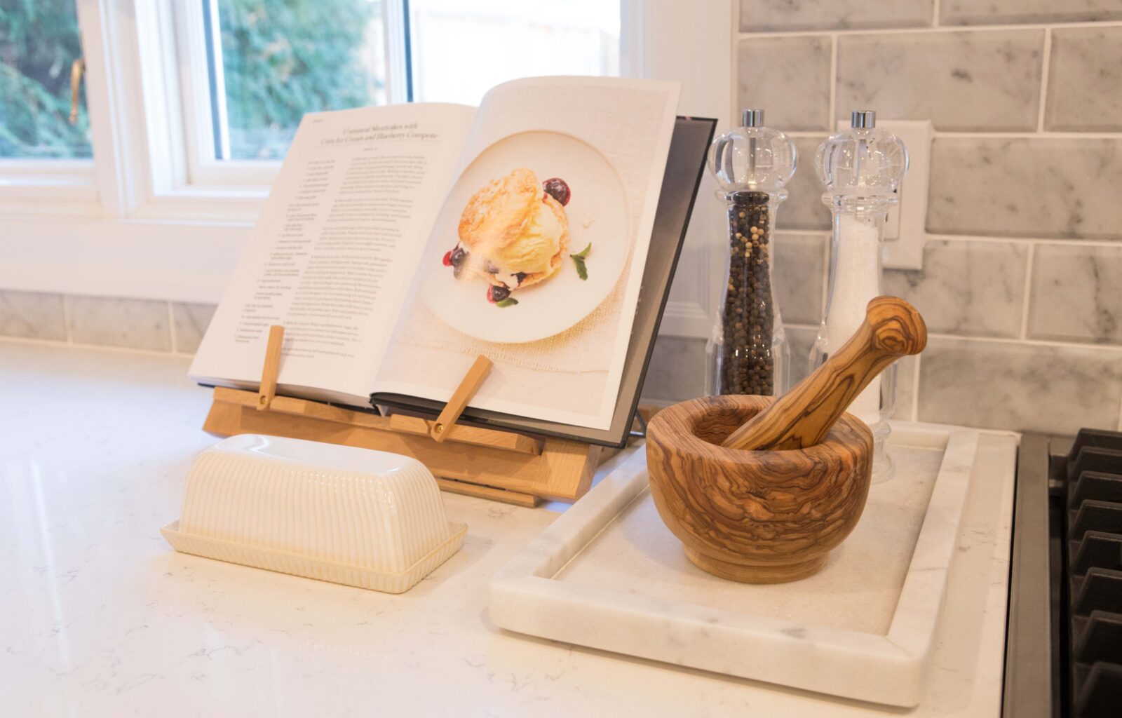 Kitchen counter with an open cookbook on a stand, a wooden mortar and pestle, salt and pepper grinders, and a butter dish