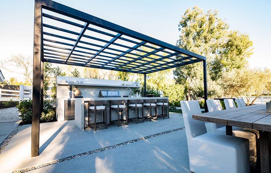 Outdoor kitchen and dining area with bar seating under a pergola and a wooden dining table with white chairs