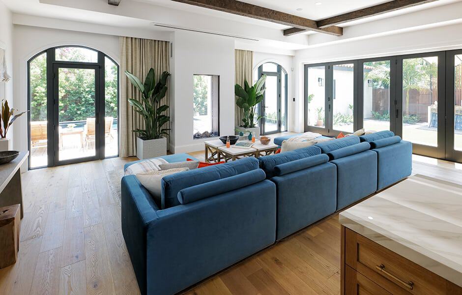 Bright living room with a large blue sectional sofa, wood floors, tall plants, and wide glass doors