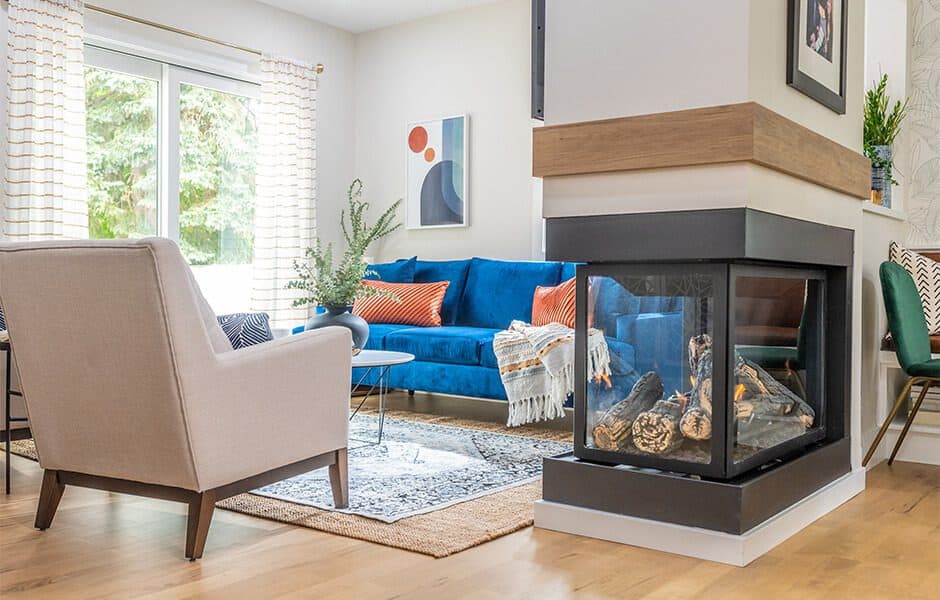Living room with blue sofa, coffee table, patterned rug, three-sided glass fireplace, windows with curtains, and a chair