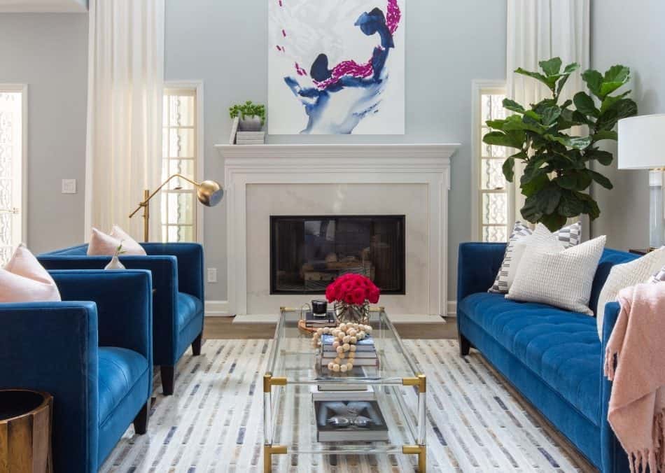 Living room with blue sofas, a glass coffee table, fireplace, large artwork, and indoor plants