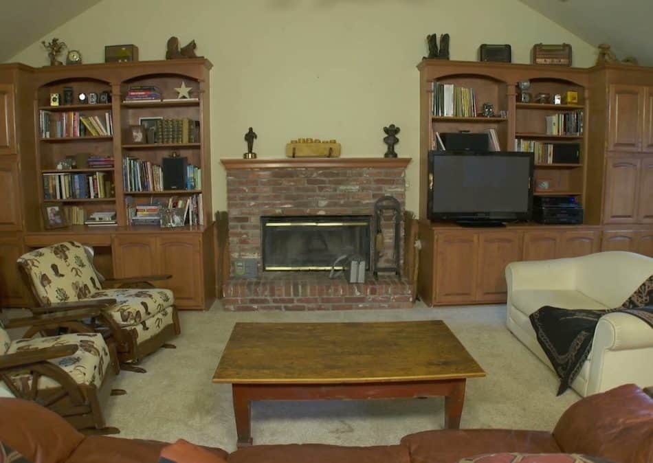 Dated living room with brick fireplace, bookshelves, coffee table, carpeted floor, ceiling fan, TV, armchairs, and sofa