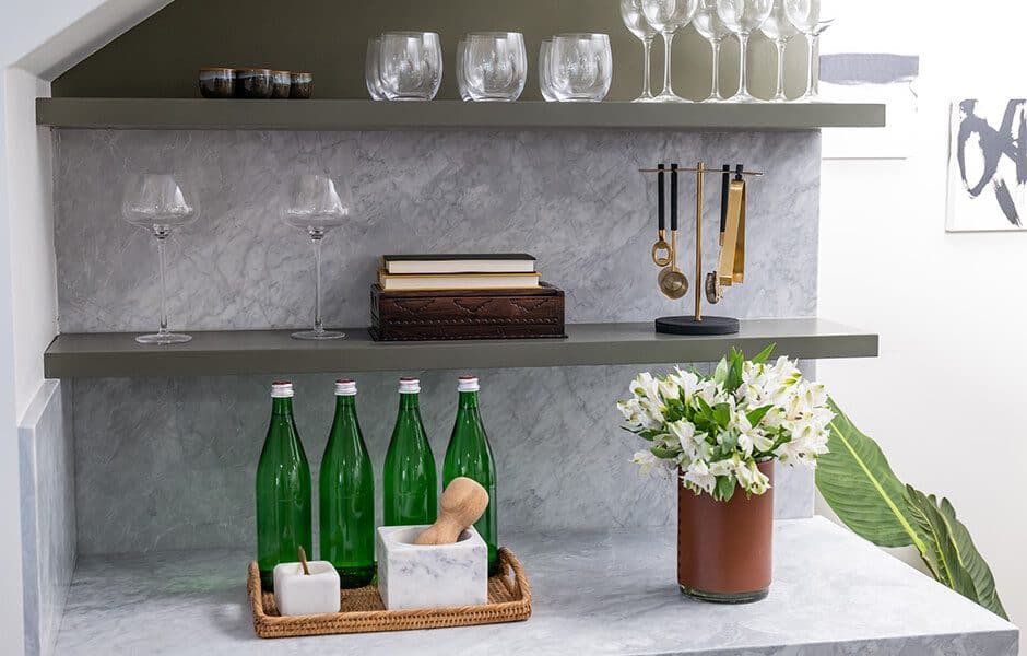 Elegant marble bar nook with glassware, green bottles, books, and a floral arrangement