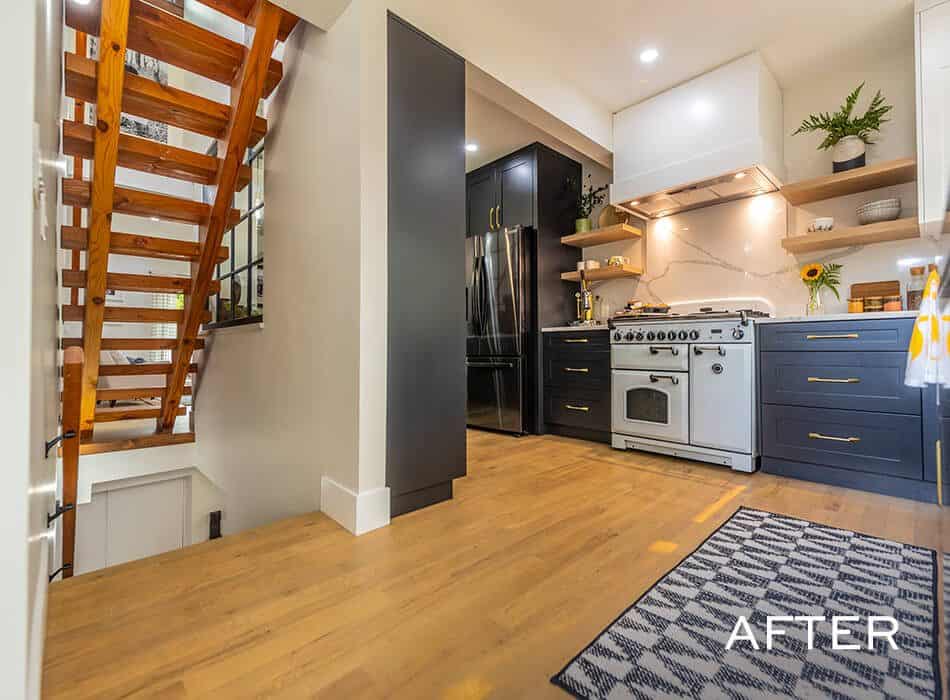A kitchen with light wood floors, black wall, white staircase, dark blue cabinets, white range, marble countertop, and floating shelves