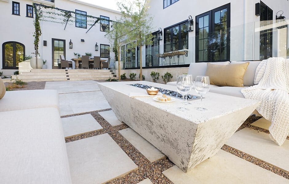 Outdoor patio with fire table, beige seating, and dining area by a white house with black windows