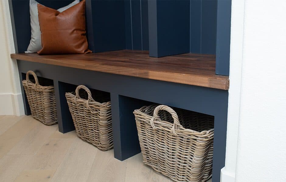 Wooden bench with navy blue base, wicker storage baskets underneath, and accent pillows on top