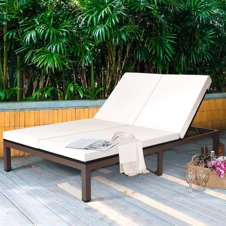 Outdoor double chaise lounge with white cushions, set on a deck beside lush green plants
