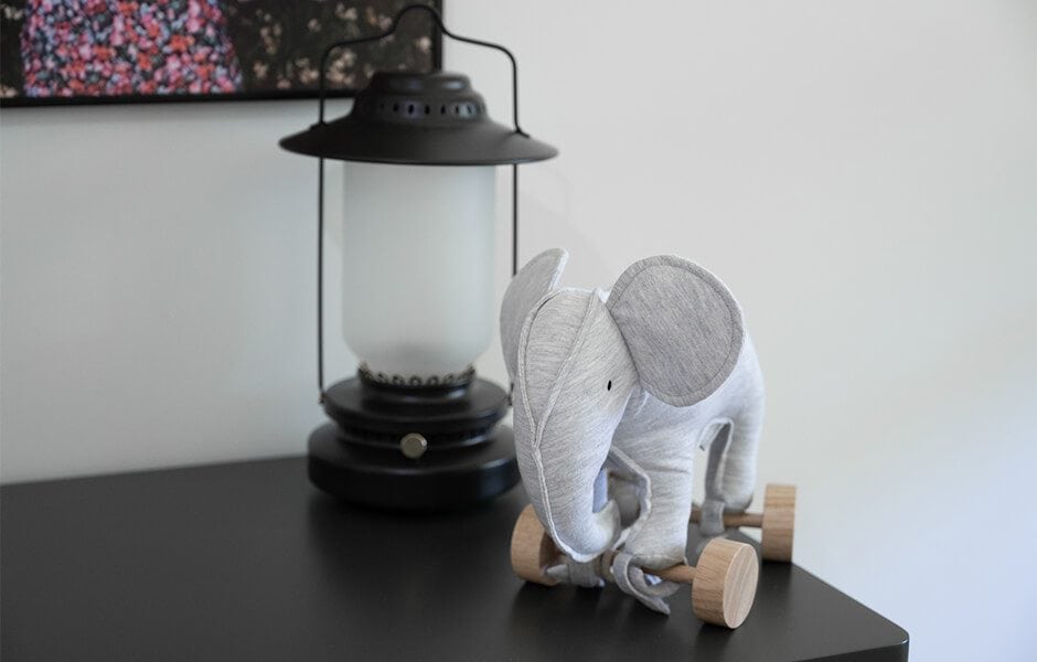 A gray stuffed elephant toy with wooden wheels sits on a table next to a black lantern