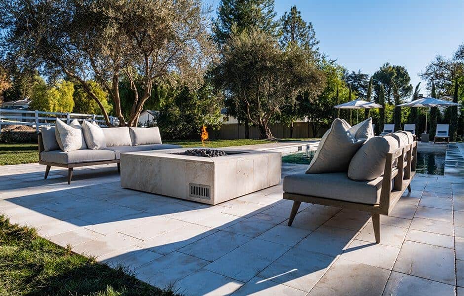Outdoor patio with cushioned seating around a square stone fire pit, set on tiled flooring beside a swimming pool