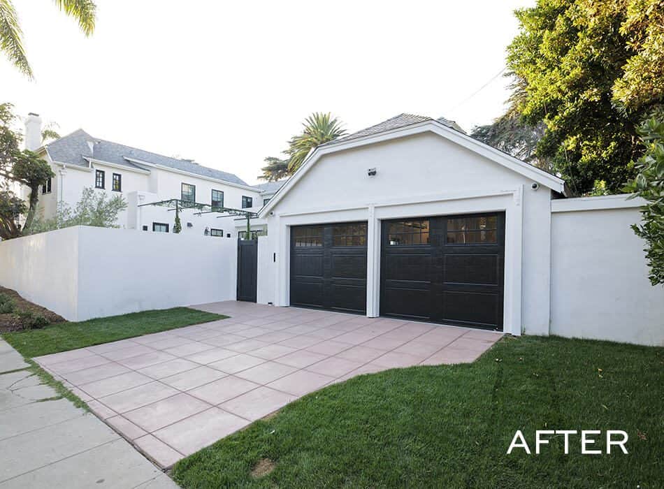 Modern white garage with two black doors, a pink-tiled driveway, and surrounding green lawn next to a large house