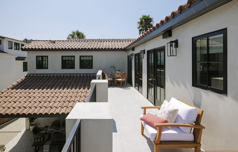 Outdoor balcony with tiled flooring, wooden seating with cushions, black-framed windows, and a terracotta roof