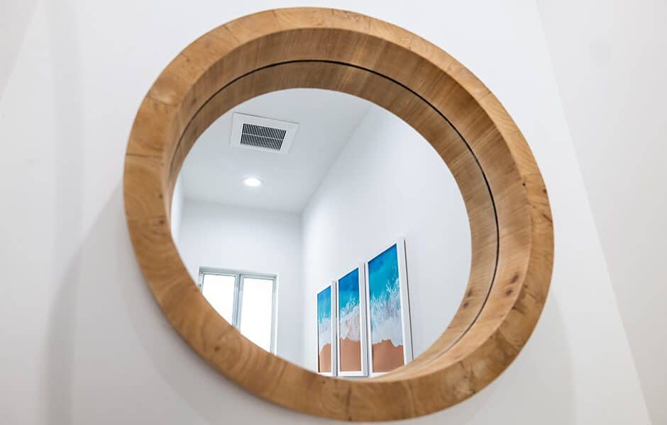 Round wall mirror with a thick wooden frame reflecting artwork and a window in a bright hallway