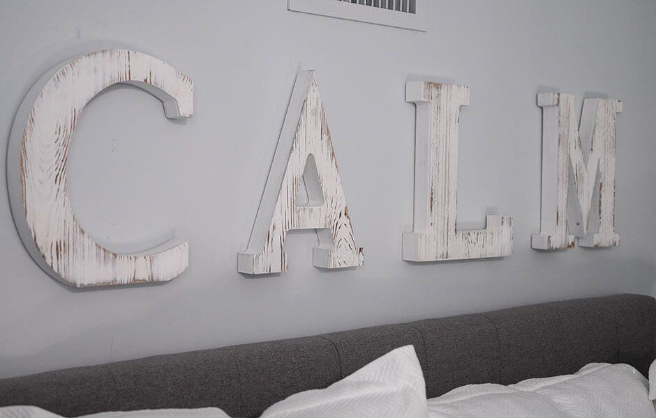 The image features the word "CALM" in whitewashed wooden letters above a gray bed