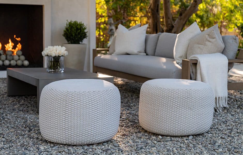 Outdoor seating area with a gray sofa, white knit poufs, a low table with flowers, and a modern fireplace on a gravel patio