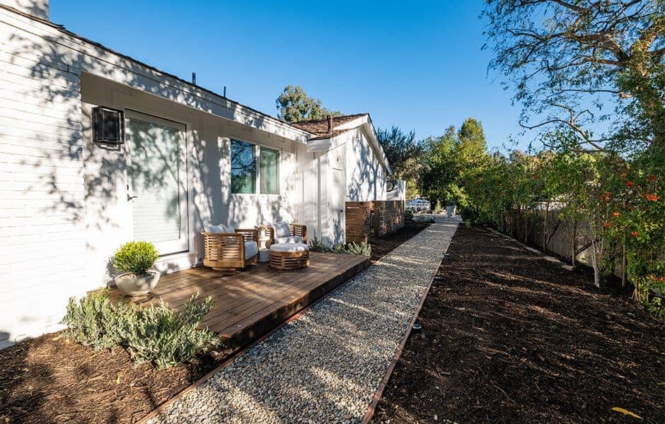 Backyard with gravel walkway, small wooden deck seating area, potted plants, and greenery along the fence