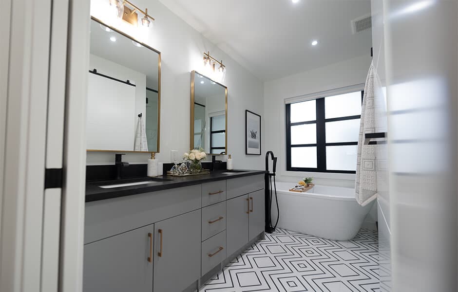 Modern bathroom with double vanity, black countertops, gold-framed mirrors, geometric tile floor, and a freestanding tub by the window