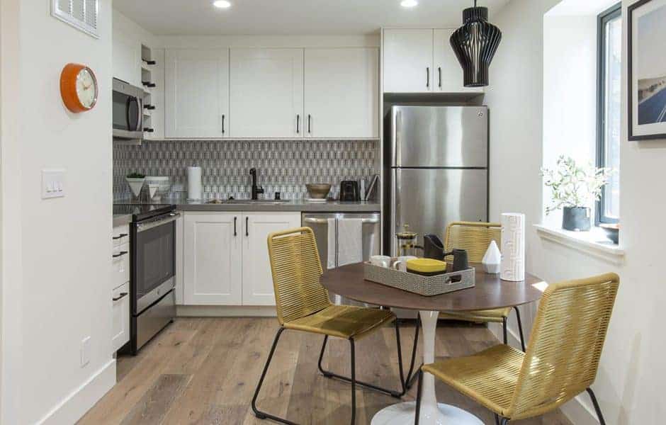 Compact kitchen with white cabinets, stainless steel appliances, patterned backsplash, and a round dining table with yellow chairs