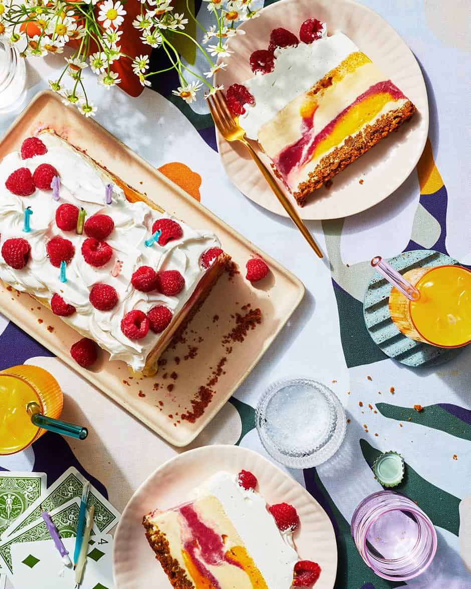 Colorful fruit and cream cake topped with raspberries, served with slices on plates alongside drinks and flowers on a patterned table