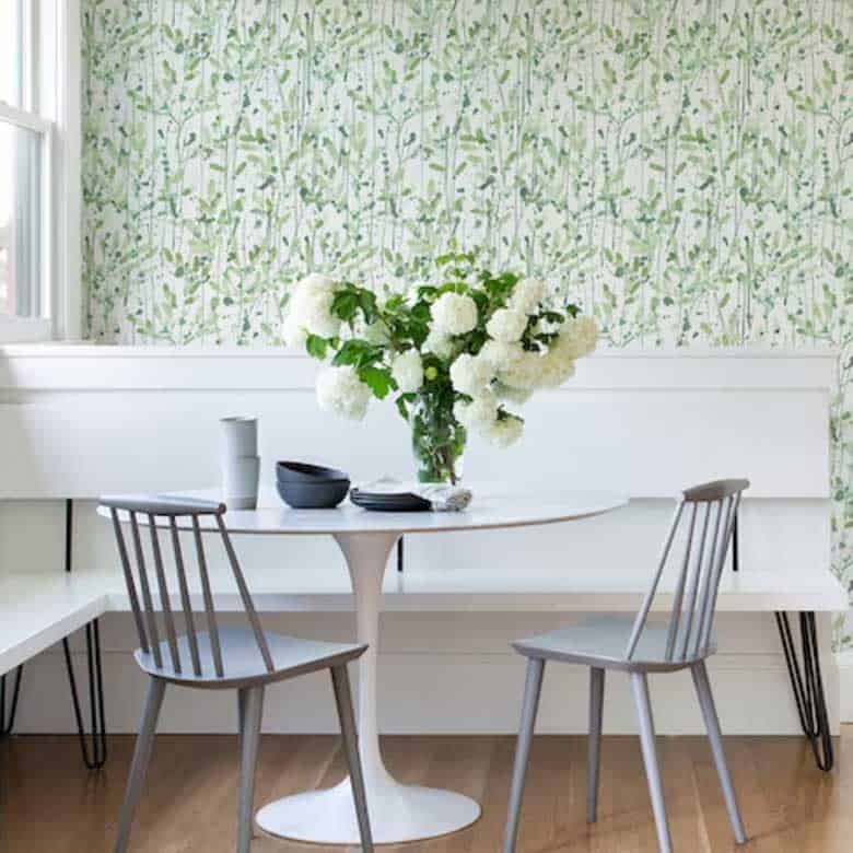 Bright dining nook with a round white table, gray chairs, and a vase of white flowers against a leafy patterned wallpaper
