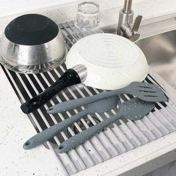 Kitchen utensils, a frying pan, and a bowl drying on a roll-up dish rack over a sink