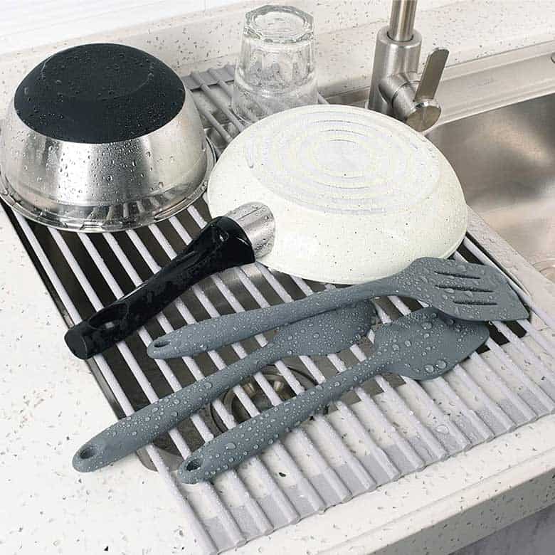 Kitchen utensils, a frying pan, and a bowl drying on a roll-up dish rack over a sink
