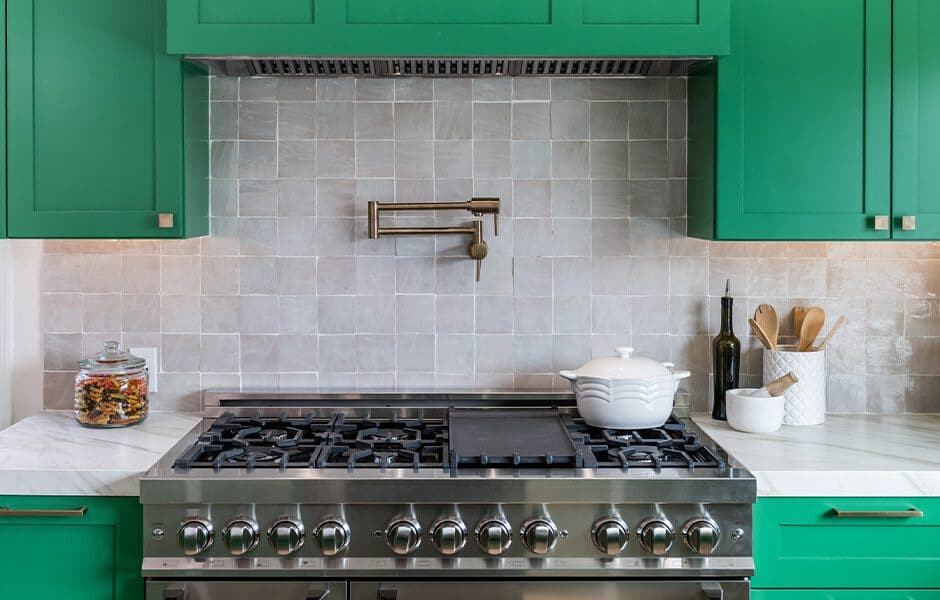 Stainless steel gas stove with a white pot, green cabinets, a tile backsplash, and a wall-mounted pot filler
