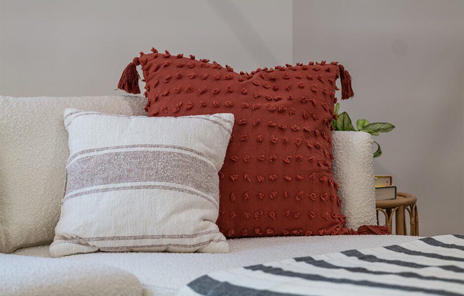 Two decorative pillows on a light-colored sofa: one textured red pillow with tassels and a smaller striped neutral pillow