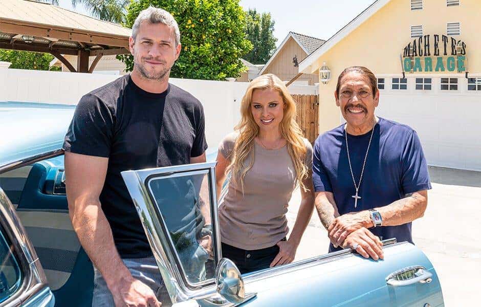 Mario, Cristy, and Danny Trejo standing beside a vintage blue car outside a garage labeled 