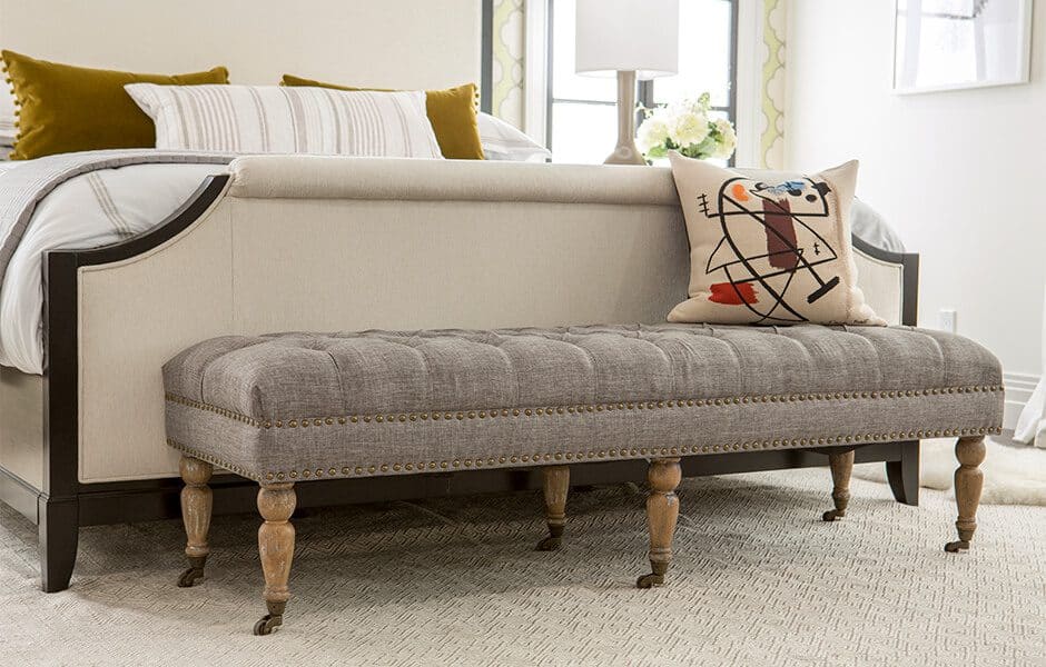 Bedroom with a tufted gray upholstered bench at the foot of the bed, decorated with a patterned pillow and neutral bedding