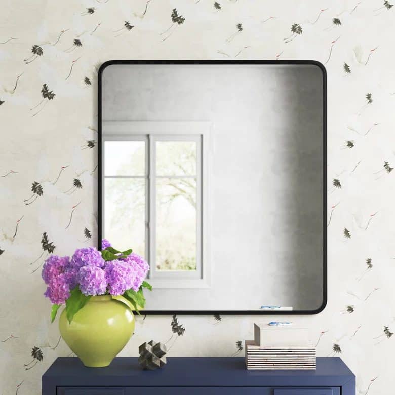 Mirror with black frame, purple flowers in a green vase, books, and a geometric object on a blue dresser