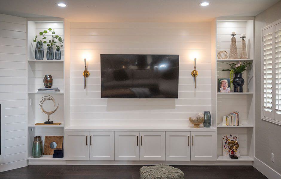 Living room entertainment wall with a mounted flat-screen TV, white shiplap backdrop, built-in shelving, accent lighting, and decorative items
