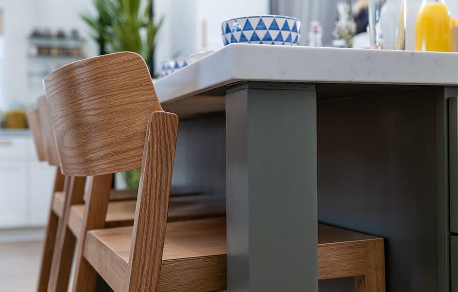 A close-up of a kitchen island with a white marble countertop and gray legs, together with wooden counter stools with curved backs