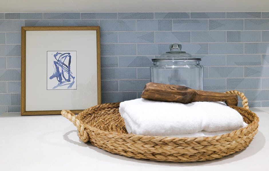 Woven basket with towels, brush, framed art, and glass jar against a blue tiled backsplash