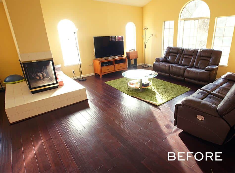 A before image of a living room with yellow walls, dark wood flooring, a fireplace, leather sofas, and a green rug