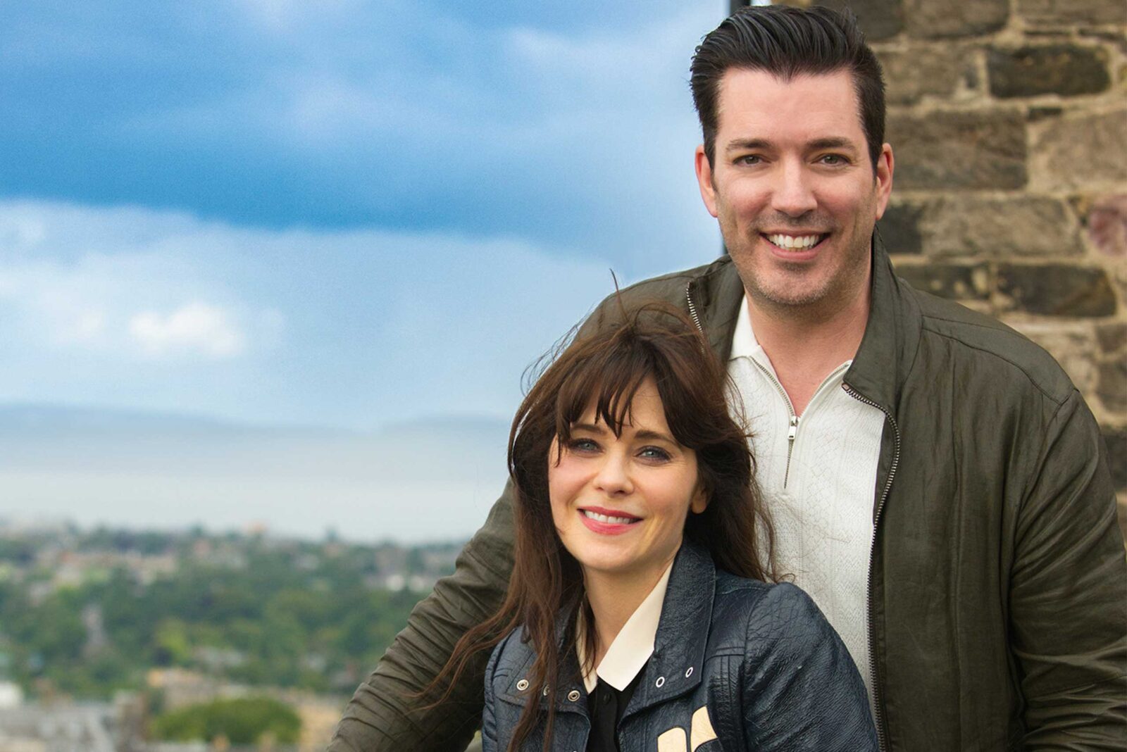 Jonathan and his wife posing outdoors with a scenic city view and stone wall in the background