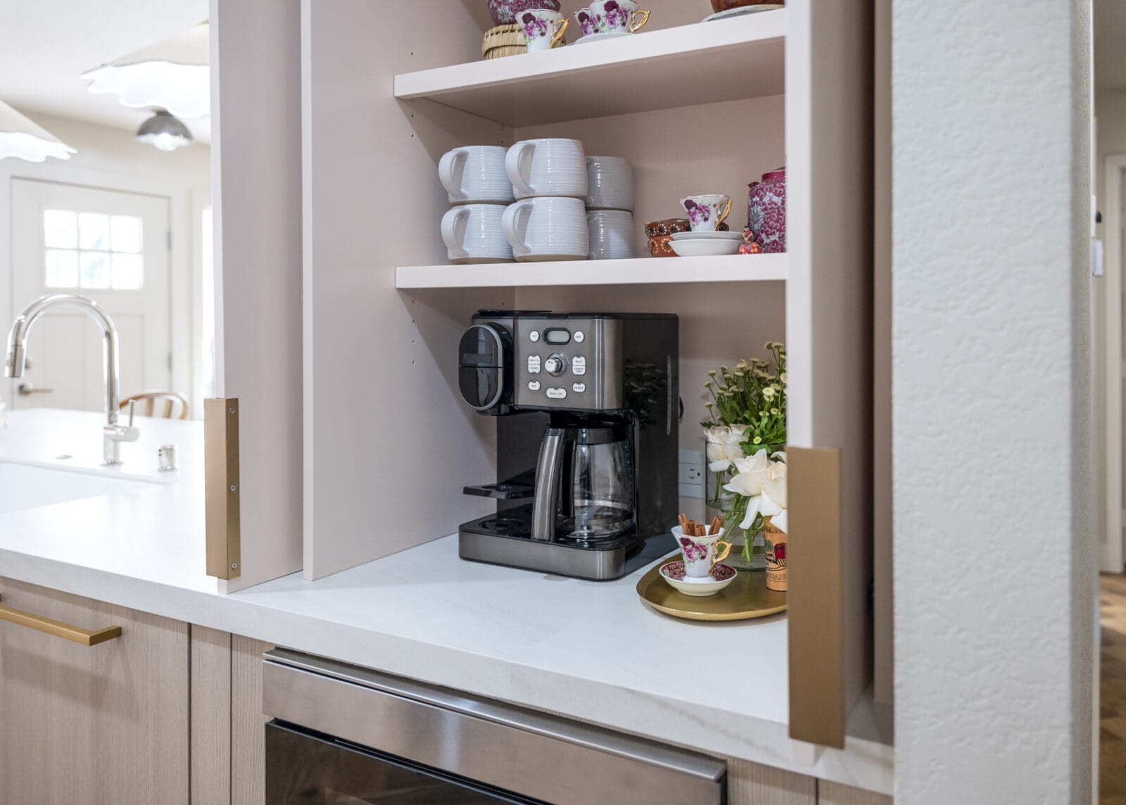 Coffee station with a black coffee maker, stacked mugs, floral teacups, and a vase of white flowers on a kitchen countertop shelf