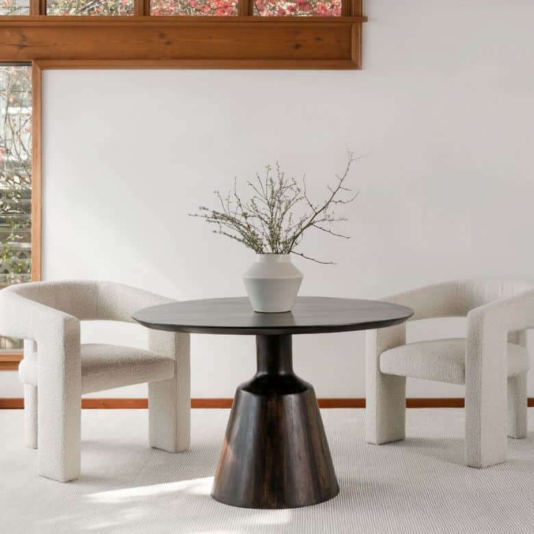 Minimalist dining area with two cream chairs, a dark round table, and a white vase with branches