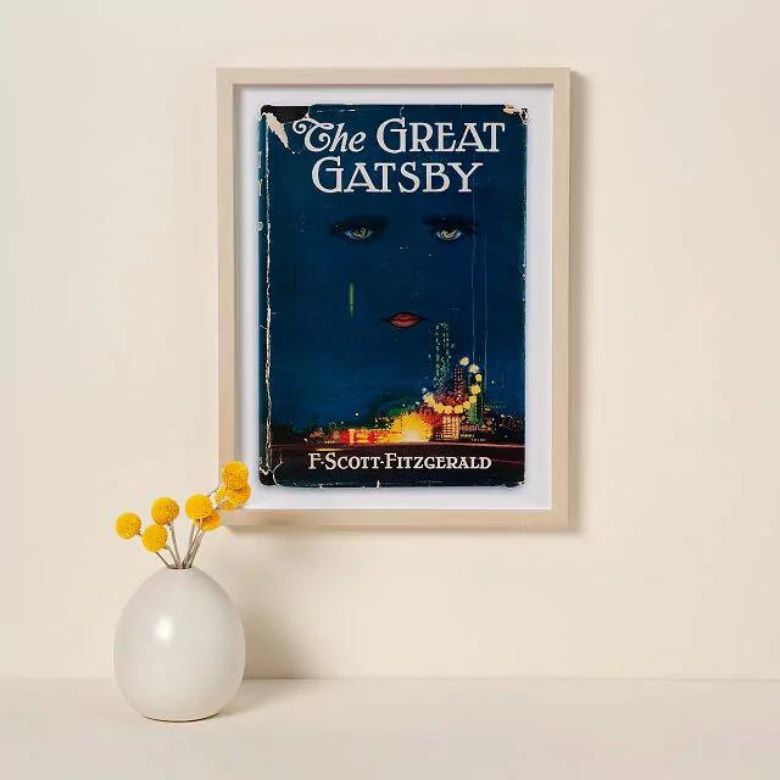 Framed “The Great Gatsby” book cover on a wall above a white vase with yellow flowers