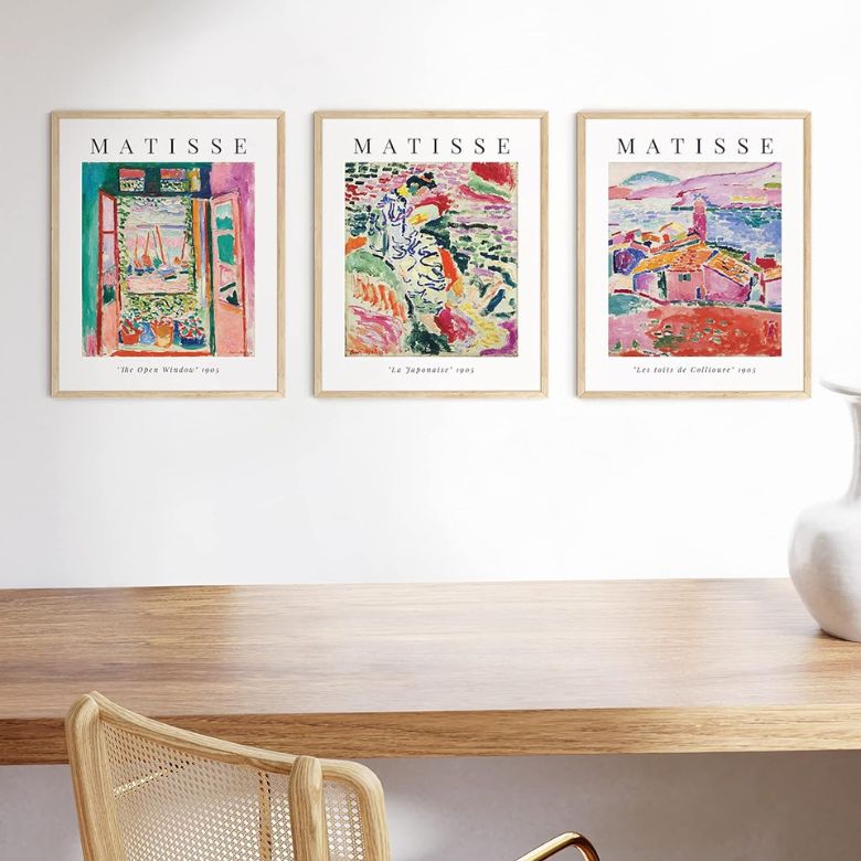 Three framed Matisse art prints hang above a wooden desk with a woven chair and a white vase in a minimalist setting