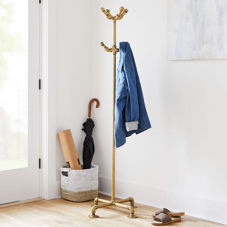 Brass coat rack stands in a corner with a blue jacket hanging, next to a basket holding an umbrella and rolled mat.